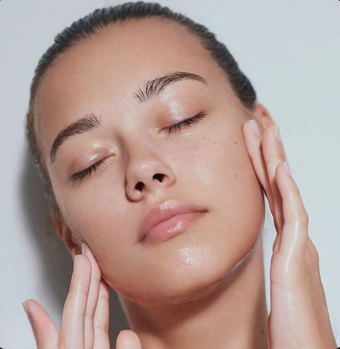 Gros plan sur le visage d'une femme aux yeux fermés, appliquant délicatement un soin hydratant et brillant sur sa peau propre.